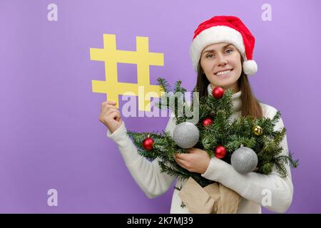 Grazioso giovane in cappello rosso di Natale che tiene il bouquet dei rami dell'abete rosso e del simbolo giallo grande del hashtag, posante isolato sopra la parete di fondo viola del pastello Foto Stock
