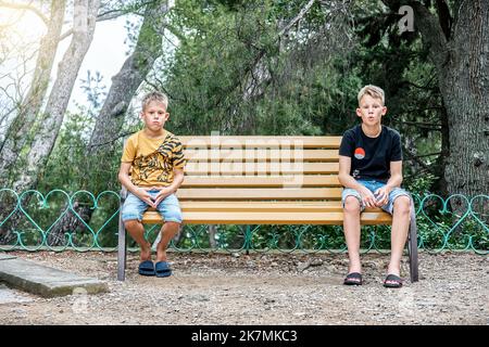 I fratelli seduti su una panca di legno guardano con espressioni sconvolgenti e deluse. I fratelli siedono su diversi lati della panchina in silenzio dopo le discussioni Foto Stock