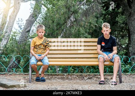 I fratelli seduti su una panca di legno guardano con espressioni sconvolgenti e deluse. I fratelli siedono su diversi lati della panchina in silenzio dopo le discussioni Foto Stock