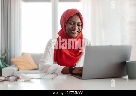 Lavoro remoto. Felice donna islamica in hijab rosso che lavora su un computer portatile, digitando sulla tastiera e guardando lo schermo del computer Foto Stock