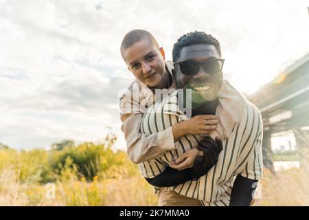 Splendido ritratto all'aperto di coppie multietniche che si godono il tempo all'aperto. Bell'alto uomo nero in occhiali da sole sorridente alla macchina fotografica, prendendo la sua ragazza calva caucasica sul piggyback. Foto di alta qualità Foto Stock