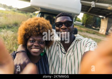 Selfie POV. Adorabile coppia nera eterosessuale che prende un selfie o fa una videochiamata, sorridendo alla fotocamera mentre si trova fuori dal loro pulmino. Foto di alta qualità Foto Stock