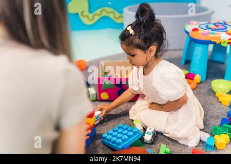 full shot di una piccola bella ragazza dai capelli scuri sul pavimento che gioca con giocattoli e blocchi da costruzione all'asilo. Foto di alta qualità Foto Stock