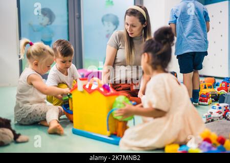 'Vogliamo costruire una città, insegnante' - i bambini caucasici adorabili divertirsi con il loro bel maestro, asilo, stanza piena di giocattoli sullo sfondo. Foto di alta qualità Foto Stock