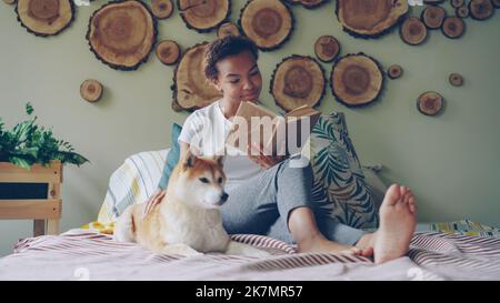 Attraente ragazza afroamericana sta leggendo il libro e accarezzando il suo adorabile cucciolo seduto a piedi nudi sul letto in appartamento moderno. Amare gli animali, godersi la letteratura e il concetto di case. Foto Stock