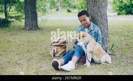 Grazioso African American ragazza giovane studente lettura libro seduto in parco sul prato, mentre il suo cane ben allevato è seduto vicino a lei e odore di aria e guardando intorno. Foto Stock