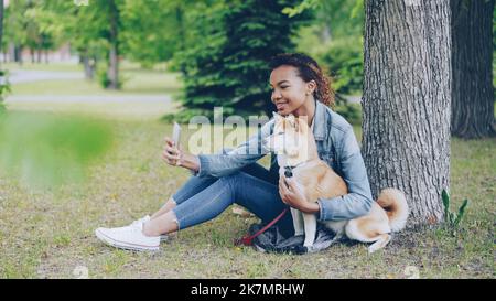 Attraente giovane ragazza afroamericana sta prendendo selfie con cane obbediente cute che riposa nel parco cittadino cocdling e carezzando l'animale bello. Tecnologia moderna, animali domestici amorevoli e concetto di natura. Foto Stock