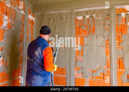 L'uomo di lavoratore sta tenendo il tubo flessibile di mortaio nella sua mano e intonacando sotto pressione muri di mattoni interni nell'edificio in costruzione. Foto Stock