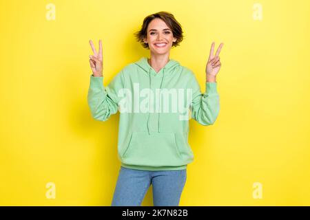 Foto di ottimista bella dolce ragazza con pettinatura di bob indossare jeans verde felpa con cappuccio che mostra segno V isolato su sfondo di colore giallo Foto Stock