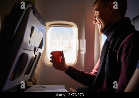 Sorridente passeggero della compagnia aerea con una tazza di caffè di carta guardando avanti nella finestra dell'aereo. L'uomo beve il tè a bordo. Il maschio trattiene bevande o acqua durante Foto Stock