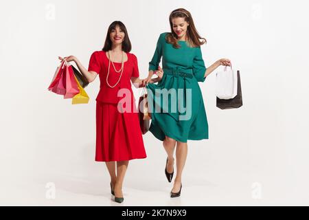 Due donna sorridente attraente con borse di shopping a colori in piedi su un bianco. Ragazze piuttosto giovani in stile retrò 70s, 80s, 90s moda, abiti isolati Foto Stock