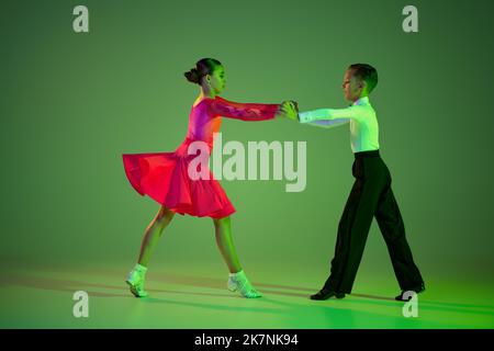 Studio shot di un grazioso bambino e bambina ballare ballo sala da ballo isolato su sfondo verde in luce al neon. Concetto di arte, bellezza, grazia, azione Foto Stock