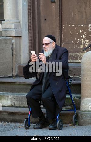 Un più anziano ebreo si siede sul suo camminatore e a sembra essere un raro uomo hassidico che usa uno smartphone. A Brooklyn, New York. Foto Stock