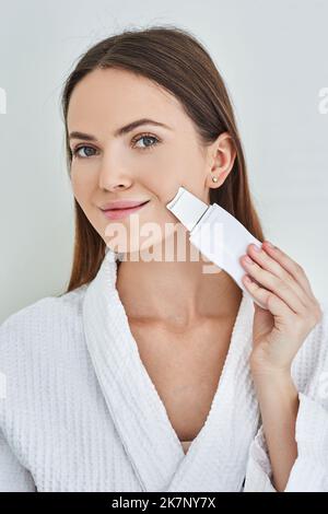 Pelatura ultrasonica del viso e del mento per una donna graziosa con scrubber ultrasonico, su sfondo grigio chiaro. Pulizia della pelle del viso, rimozione delle teste nere Foto Stock