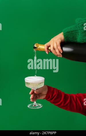 Sfondo astratto della festa di Natale, lo Champagne viene versato da una bottiglia in un bicchiere in una mano femminile. Minimalismo, sfondo verde, Natale A. Foto Stock