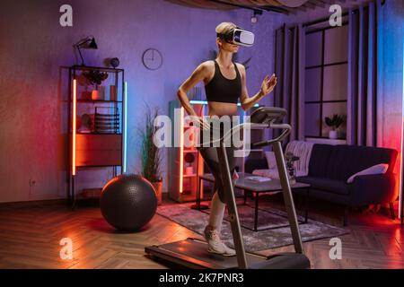 Attraente giovane sport donna caucasica fare esercizio di fitness, in esecuzione su tapis roulant utilizzando occhiali di realtà virtuale. Femminile atletica e muscolare che si allenano attivamente in interni durante la notte. Foto Stock