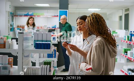 Un dipendente medico aiuta il gruppo multietnico di clienti a comprare farmaci e vitamine in farmacia, parlando di pillole sugli scaffali. Fornire assistenza con prodotti farmaceutici e farmaci. Foto Stock