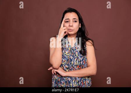 Donna indiana dubbia guardando lateralmente ritratto, modello di pensiero, prendere la decisione, tenendo il dito sulla faccia. Persona premurosa in piedi con un'espressione facciale penosa, vista frontale, scatto medio da studio Foto Stock