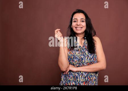 Donna indiana sorridente in posa ritratto, donna felice tenendo la mano piegata, scattando le dita. Persona allegra in piedi, guardando la macchina fotografica, vista frontale studio medio girato su sfondo marrone Foto Stock