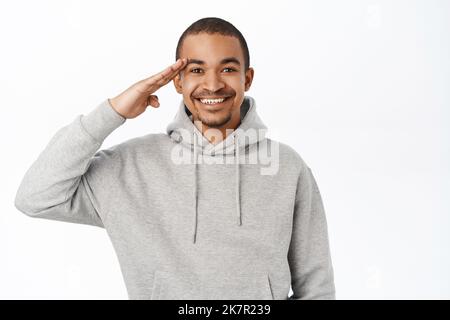 Immagine del giovane uomo che saluta, tiene la mano vicino alla fronte, saluto dell'esercito, in piedi su sfondo bianco Foto Stock