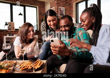 Giovane coppia afro-americana che mostra le foto sullo schermo del telefono delle cellule alle donne differenti di etnie, tutto sorride. Amici di gruppo multiculturali seduti, in movimento, sorridenti, parlanti, in discussione. Foto Stock
