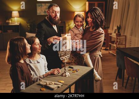 Ritratto di famiglia ebraica moderna illuminazione in argento menorah candela durante la celebrazione Hanukkah in accogliente ambiente casa Foto Stock