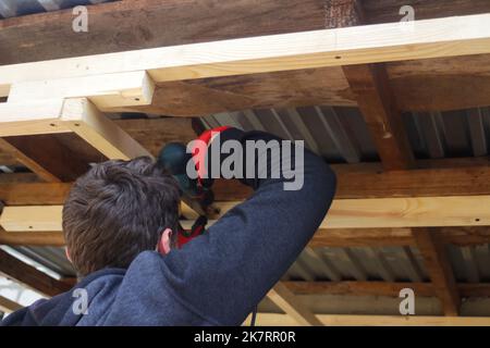Defocalizzare l'operatore utilizzando la seminatrice che lavora sulla costruzione di case e tetto con telaio in legno. Uomini che fissano la mensola di legno con il suo Driller. Fuori fuoco Foto Stock