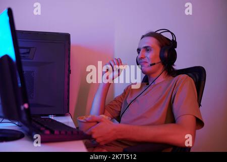 Giovane uomo sorridente che mangia patatine fritte guardando il flusso dal vivo del blogger preferito sullo schermo del computer Foto Stock