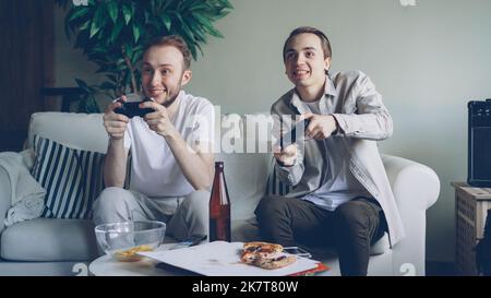 Due bei ragazzi stanno giocando a un videogioco con joystick seduti sul divano di casa. Gli uomini emotivi stanno godendo di gioco, spuntini e bottiglie di birra sono visibili. Foto Stock