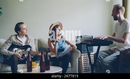 Una bella cantante bionda sta cantando e ridendo durante le prove con la sua band musicale a casa. Il ragazzo bearded sta suonando le tastiere e il giovane allegro sta suonando la chitarra. Foto Stock