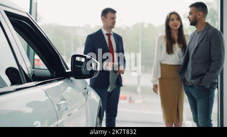 Il venditore di automobile professionale nel vestito sta avendo conversazione con la coppia ricca attraente che dice loro circa il nuovo modello dell'automobile, i compratori allegri stanno sorridendo. Messa a fuoco su bello lucido auto. Foto Stock