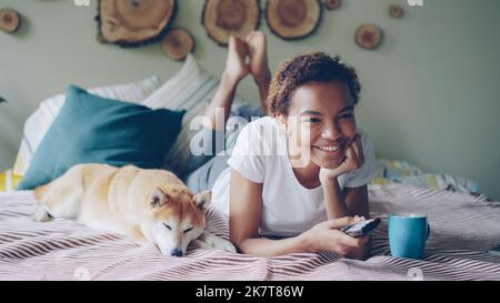 Giovane afroamericana sta guardando la TV che tiene il telecomando premendo i tasti e ridendo mentre il suo cane da compagnia è sdraiato accanto a lei. Concetto di intrattenimento e tecnologia. Foto Stock