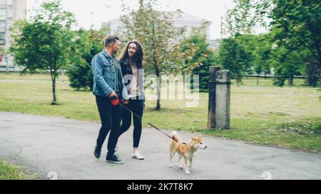 Ragazzo allegro in abiti denim è a piedi il suo cane tenendo la mano di sua moglie, la gente e l'animale stanno andando lungo il percorso nel parco in estate. Stile di vita attivo e concetto di animali domestici. Foto Stock