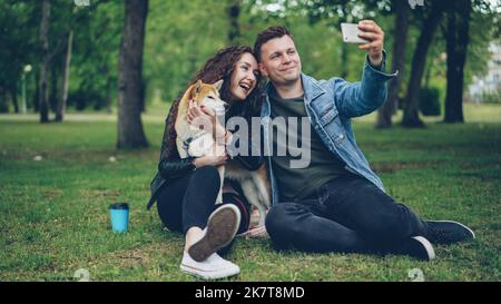 Bel giovane uomo sta prendendo selfie con la sua ragazza che tiene il cane pedigree, la gente sta ridendo. Foto Stock