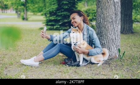 Attraente giovane ragazza afroamericana sta prendendo selfie con cane obbediente cute che riposa nel parco cittadino cocdling e carezzando l'animale bello. Tecnologia moderna, animali domestici amorevoli e concetto di natura. Foto Stock