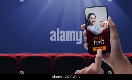 Donna che tiene uno smartphone e acquista biglietti per film online utilizzando un'app mobile, una sala cinema e posti a sedere in background, ripresa POV Foto Stock