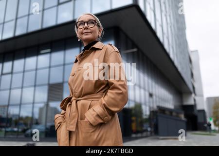 una donna anziana di successo si trova sullo sfondo della facciata di un moderno edificio di uffici Foto Stock