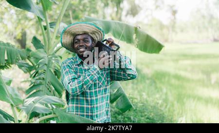 L'uomo contadino africano ascolta il ricevitore radio retrò sulla spalla sorride felice nella sua fattoria biologica Foto Stock
