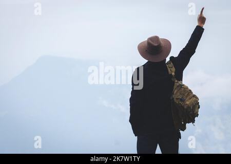 Un viaggiatore maschio in uno zaino si trova in cima alla montagna con i pollici in alto che mostrano il segno del "numero uno" in una giornata di nebbia e pioggia. Avventur Foto Stock