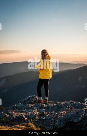 Donna viaggiatore beve caffè dopo una camminata riuscita alla cima della montagna. Escursionista femminile godendo il tramonto sulla catena montuosa. Avventura nella natura Foto Stock