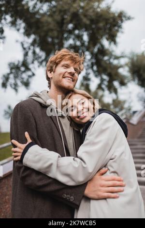felice giovane donna che abbraccia il ragazzo rosso testa in cappotto autunnale in parco, immagine stock Foto Stock