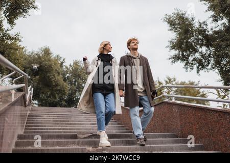 vista dall'angolo basso di una donna allegra e di un uomo rosso con ombrello che tiene le mani mentre scendi le scale vicino al parco, immagine stock Foto Stock