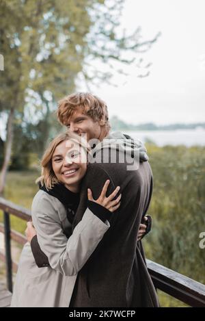 giovane donna gioiosa sorridente e abbracciante uomo rosso durante la data sul ponte vicino stagno, immagine stock Foto Stock