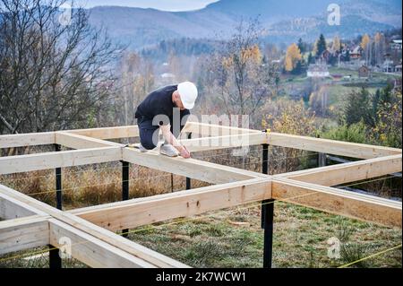 Uomo lavoratore costruire casa telaio in legno su fondazione palo. Falegname che misura tavole di legno e facendo segni con matita. Concetto di Falegnameria. Foto Stock