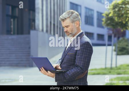 boss dai capelli grigi seri e concentrati che lavora sul notebook mentre si trova fuori dall'edificio degli uffici moderni, boss maturo in tuta aziendale che digita sulla tastiera del netbook, uomo d'affari di successo senior. Foto Stock