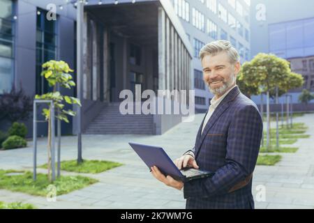 Ritratto di un uomo d'affari maturo fuori dal moderno edificio degli uffici, investitore senior dai capelli grigi con notebook sorridente e guardando la fotocamera, uomo di successo in tuta aziendale con netbook. Foto Stock