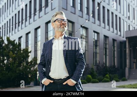 Ritratto di investitore senior di successo, business maturo fuori ufficio edificio in tuta e occhiali da lavoro, grigio haired banchiere sorridendo e guardando via. Foto Stock