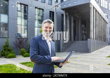 Ritratto di un uomo d'affari maturo fuori dal moderno edificio degli uffici, investitore senior dai capelli grigi con notebook sorridente e guardando la fotocamera, uomo di successo in tuta aziendale con netbook. Foto Stock