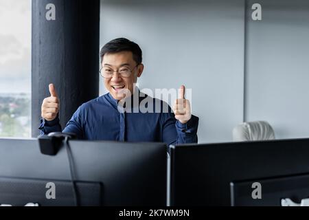 Foto ravvicinata, felice uomo d'affari asiatico che celebra la vittoria, uomo che guarda lo schermo del computer e tiene i pollici in su gioiosamente e affermativamente, il mediatore trionfa sulla borsa. Foto Stock