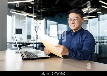 Felice uomo asiatico in occhiali ricevuti lettera apre e legge il messaggio documento, l'uomo d'affari lavora all'interno di un moderno edificio ufficio sul computer portatile, buone notizie nella notifica. Foto Stock